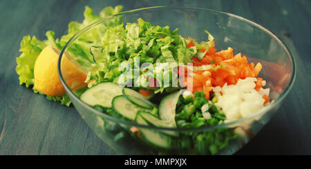 Glass bowl with cut vegetables for a salad. Close up. Bell pepper, onion, leafy greens and cucumbers cutted in pieces. Wooden background. Stock Photo