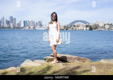 Young woman stands on the background of the beautiful Sydney skyline on a sunny day in a white dress and sunglasses Stock Photo