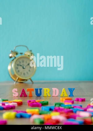 Colorful wooden word SATURDAY on wooden table and vintage alarm clock and background is powder blue. English alphabet made of wooden letter color. Stock Photo