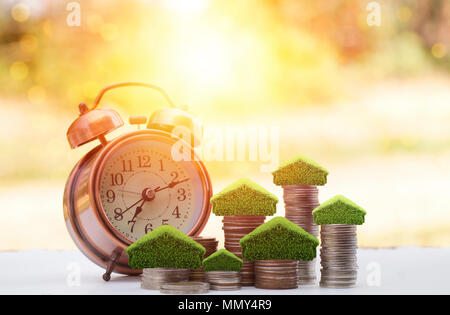 money on stacked of coins to save for house, small tree and home on pile with alarm clock on wood table with sunlight background, concept as investmen Stock Photo