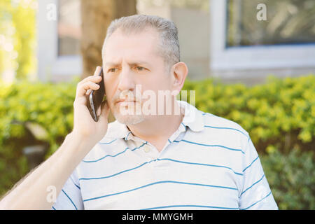 Worried middle age man talking on the phone in a park. Outdoors. Technology concept. Stock Photo