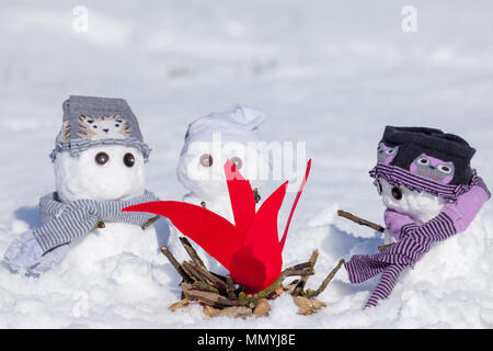 Cute little snowmen dressed for winter, huddled around a camp fire trying to keep warm. Snow covered scene in Norfolk UK. Snowman wearing clothes. Stock Photo