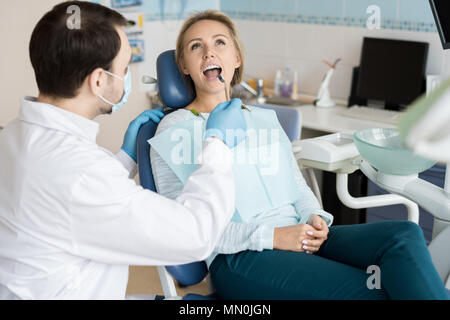 Doctor in gloves and mask exploring teeth of young girl with small mirror while sitting in chair. Stock Photo