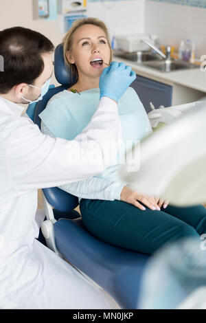Doctor in uniform and gloves using small mirror tool and checking teeth of young woman on chair. Stock Photo