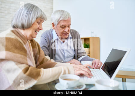 Side view of elderly man and woman using laptop sitting at table in assisted living facility and drinking tea. Stock Photo