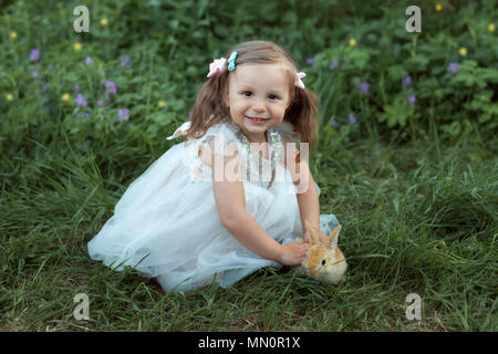 Little girl in white dress sits on grass and holds a rabbit in hands Stock Photo