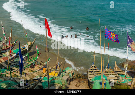 Cape Coast, Ghana - February 15, 2014: Colorful moored wooden fishing boats in African harbor town Cape Coast and locals swimming in sea Stock Photo