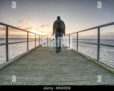 Hurt man with hooded jacket and forearm crutches  looking sadly into sea water. Traveler  stand on sea bridge within  morning and thinking. Nostalgic  Stock Photo