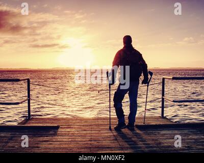 Hurt man with hooded jacket and forearm crutches  looking sadly into sea water. Traveler  stand on sea bridge within  morning and thinking. Nostalgic  Stock Photo