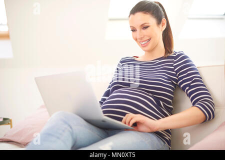 Smiling pregnant woman using laptop and browsing on the internet while sitting on sofa at home. Stock Photo