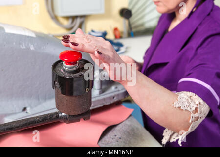 A young woman working as a seamstress in a purple unifrome presses the large red button on the industrial machine at the factory Stock Photo