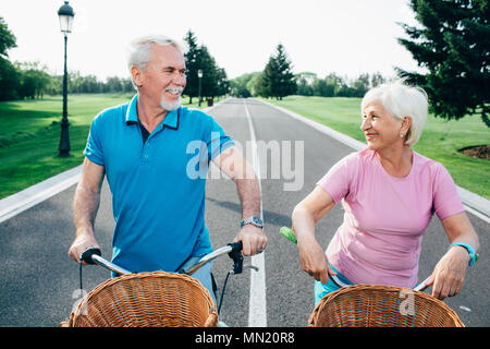 Older couple riding bicycles with baskets, standing in the middle of the road and looking into each other's eyes Stock Photo