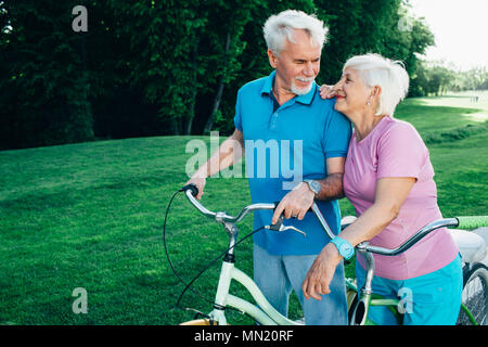 Mature couple enjoying day out on their bicycles. elderly woman hugs her husband while active rest in the park Stock Photo