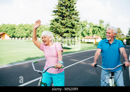 Positive senior couple bike riding in sunny park. elderly woman actively gesticulates while cycling Stock Photo