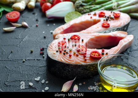 Two frash raw salmon steaks with asparagus, spinach, garlic, tomatoes, ginger, pepper, olive oil, salt, chili, onion and lemon on black background. He Stock Photo