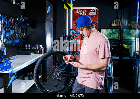 A young, stylish redheaded male small business owner selling and repairing a bike is wearing a blue cap and a pink jersey is using a mobile phone whil Stock Photo