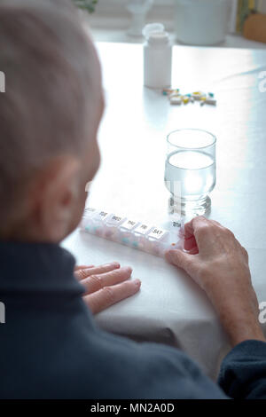 Old woman sitting at a table at home with a glass of water and lots of capsules in different colors in a presorted weekly pill dispenser. Stock Photo