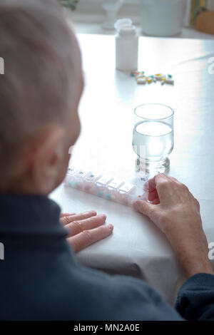 Old woman sitting at a table at home with a glass of water and lots of capsules in a presorted weekly pill dispenser GERMAN LANGUAGE Stock Photo