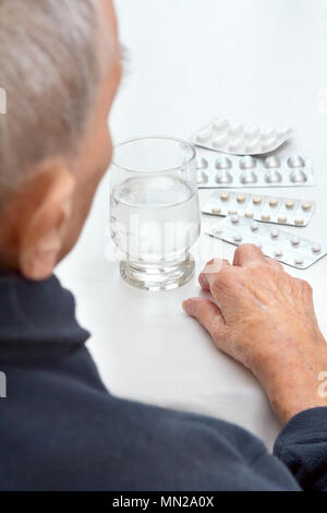 Old woman sitting at a table in her kitchen or living room with a glass of water and different tablets in blister packs Stock Photo