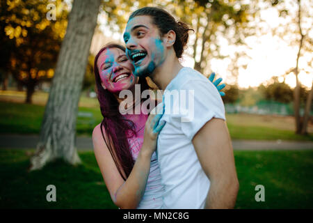 Smiling young couple with colored powder smeared on their faces. Cheerful man and woman enjoying festival of colors outdoors at park. Stock Photo