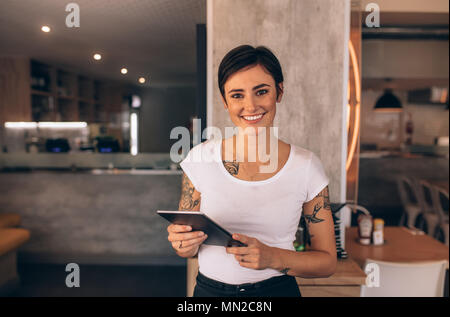 Portrait of a smiling coffee shop owner standing inside her shop. Female restaurant owner standing with a digital tablet. Stock Photo