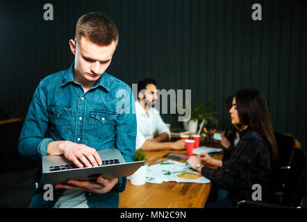 Freelancer man using laptop as standing in the office, achiving success as working on the task, busy colleagues planning project stages at brown office desk Stock Photo