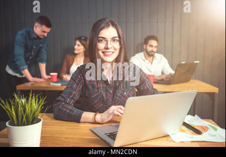 Portrait of brunette girl wears eyeglasses using silver laptop computer before office colleagues, indoor shot of working atmosphere Stock Photo