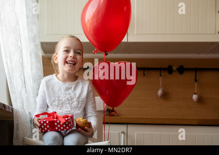 Cute preschooler girl celebrating 6th birthday. Girl holding her birthday cupcake and beautifully wrapped present, sitting surrounded by balloons. Stock Photo