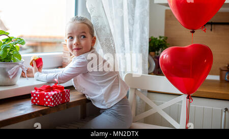 Cute preschooler girl celebrating 6th birthday. Girl with cheeky smile eating her birthday cupcake in the kitchen, surrounded by balloons. Stock Photo