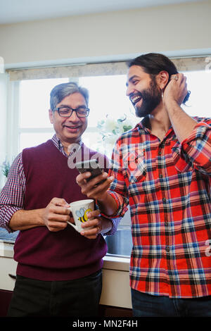 Mid adult man is showing his mature father something on his smart phone. He is holding a mug of tea. Stock Photo