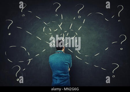 Rear view of a puzzled businessman in front of a blackboard try to solve different tasks, having questions. Stock Photo