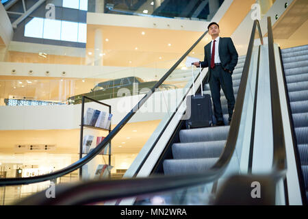 Full length wide angle portrait of successful Asian businessman descending on escalator holding suitcase in modern airport, copy space Stock Photo