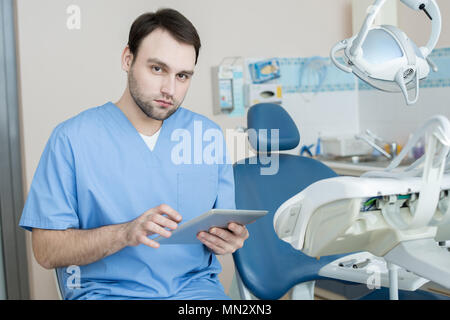 Portrait of young professional dentist posing in office by dental chair looking at camera  and using digital tablet, copy space Stock Photo