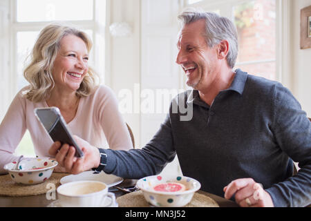 Laughing mature couple using smart phone and eating breakfast Stock Photo