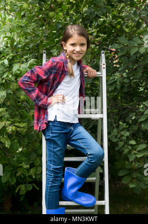 Portrait of beautiful girl in red checkered shirt and wellington boots on stepladder at garden Stock Photo