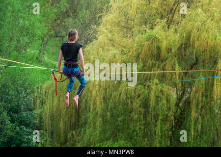 Back view of a young female tightrope walker sitting on a slackline in heights looking ahead in a park. Stock Photo