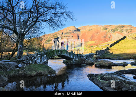 Father and children walking over Slater Bridge across the River Brathay against a backdrop of Lingmoor Fell, Little Langdale, Lake District, UK