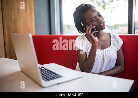 Portrait of African American girl sitting at cafe and working on her laptop talking on her cellphone while thoughtfully looking aside Stock Photo