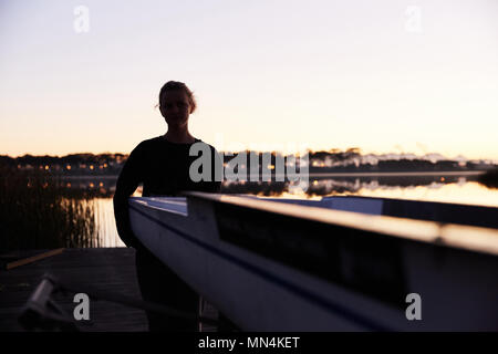 Silhouette of female rower lifting scull on sunrise lakeside dock Stock Photo