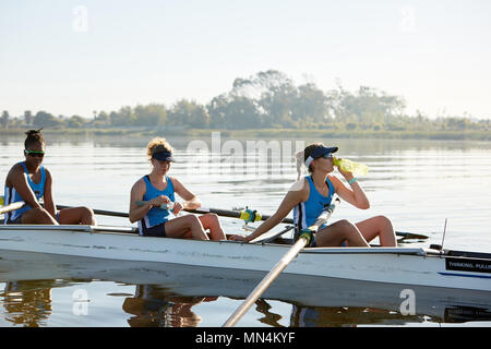 Female rowing team resting, drinking water in scull on sunny lake Stock Photo