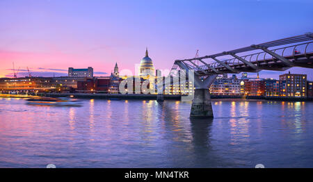 London at sunset, Millennium bridge leading towards illuminated St. Paul cathedral over Thames river with city bathing in electric light. Panoramic to Stock Photo