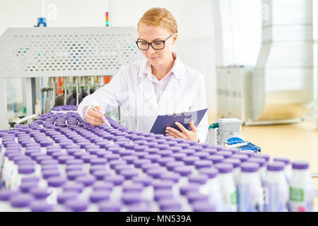 Pretty technologist wearing eyeglasses and white coat checking quality of drinking yogurts while standing at conveyor belt of dairy factory, portrait  Stock Photo