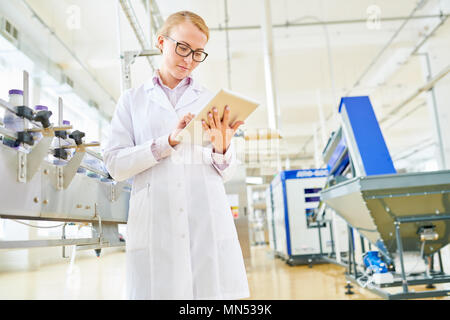 Portrait shot of pretty blonde technologist wearing white coat taking necessary notes on digital tablet while working at spacious production departmen Stock Photo
