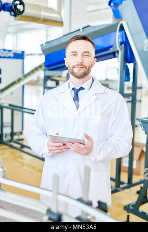Portrait shot of handsome owner of modern dairy factory wearing white coat holding digital tablet in hands while looking at camera, interior of packag Stock Photo