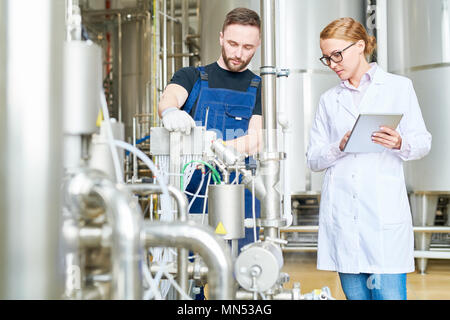 Handsome bearded technician wearing overall wrapped up in adjusting modern equipment while his female superior keeping eye on him Stock Photo