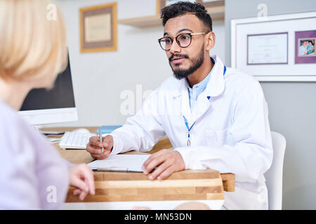 Portrait of handsome Middle-Eastern doctor consulting young woman sitting at desk in modern office, copy space Stock Photo