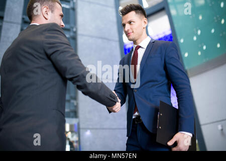 Portrait of handsome young businessman shaking hands with partner standing in hall of modern office building after closing beneficial deal Stock Photo