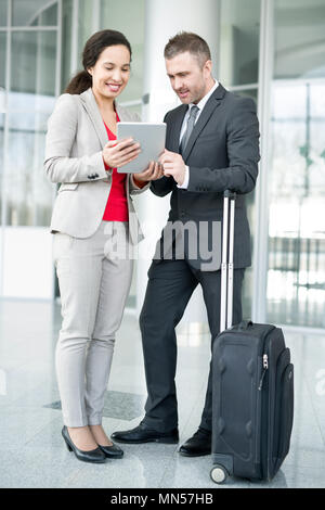 Full length portrait of successful business couple, man and woman, standing in hall of modern airport with suitcase and using digital tablet Stock Photo