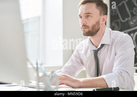 Portrait of thoughtful handsome businessman sitting at desk in office and using modern computer for work, copy space Stock Photo