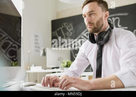 Portrait of handsome bearded businessman wearing shirt and tie sitting at desk in office and using modern computer, typing with headphones on his neck Stock Photo